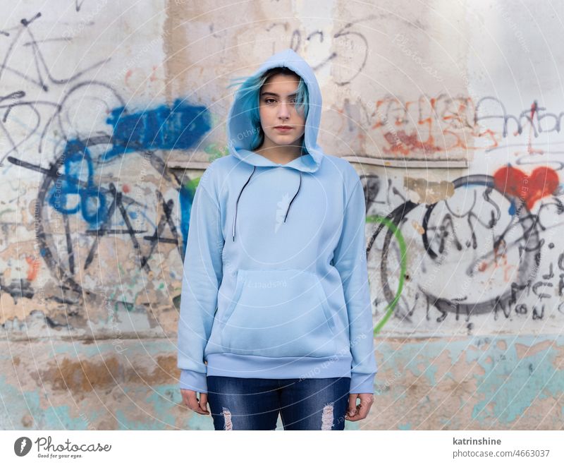 Blue haired Teenage girl in light blue oversize hoodie pointing against graffiti wall Teenager mockup jeans blue haired teen girl outdoors urban modern hipster