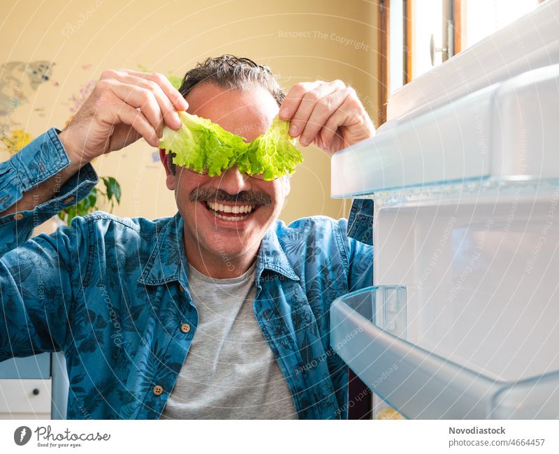 Portrait of a middle age man covering his eyes with two pieces of lettuce, with the fridge door open, and with a funny face positive optimistic middle aged