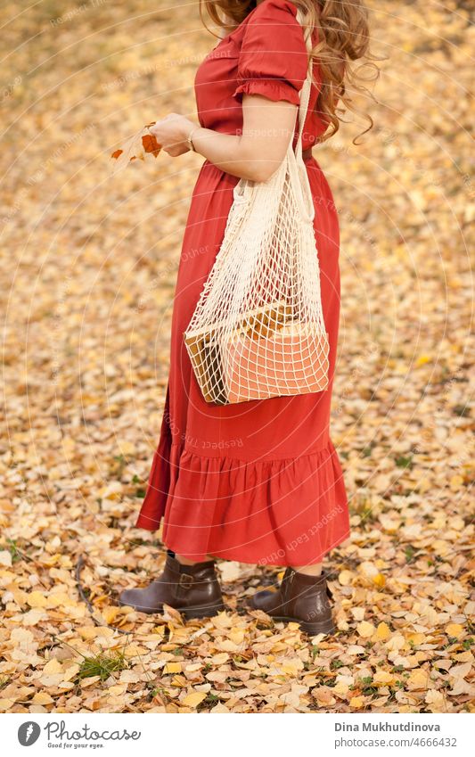 Beautiful woman in autumn park wearing a red terracota color dress, standing on autumn leaves with eco net bag with books. Young millennial woman with long hair in stylish fall outfit, looking to the camera. Autumn female lifestyle, inspiration.