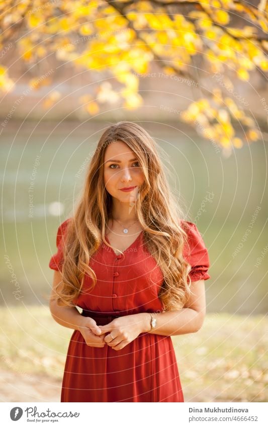 Beautiful woman in autumn park wearing a red terracota color dress, standing in autumn park near lake with yellow foliage behind her. Young millennial woman with long hair in stylish fall outfit, looking to the camera. Autumn female lifestyle, inspiration.