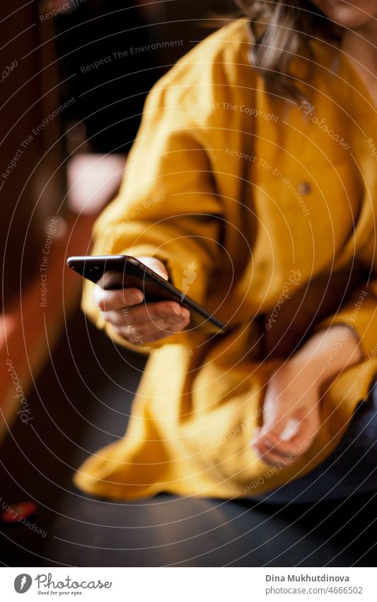Black mobile phone in female hand closeup. Unrecognizable woman in yellow clothes sitting in cafe and texting on cellphone. Real people using mobile phones in public places. Millennial and generation Z digital lifestyle.