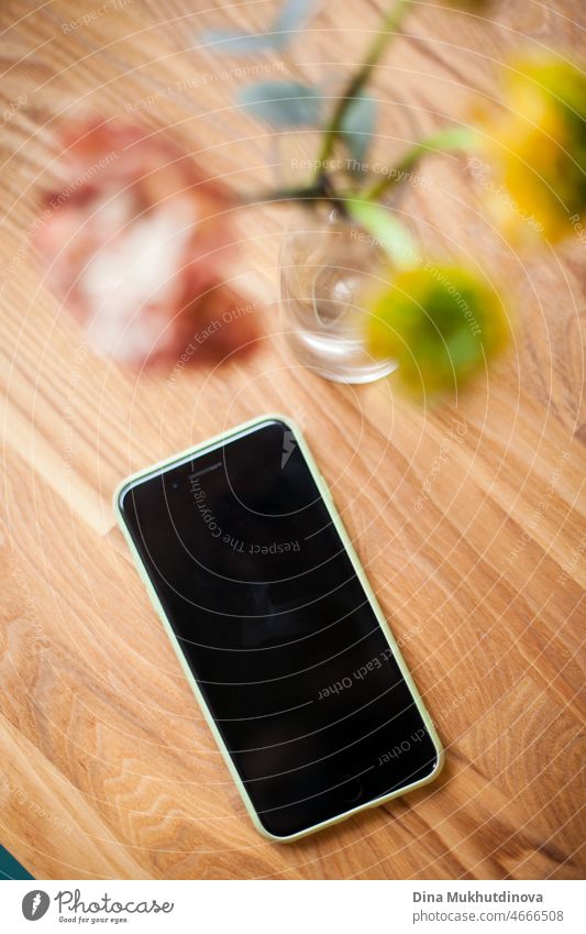 Minimalist flowers in a vase on wooden table and mobile phone - cafe lifestyle background with room for text and copy space and mockup. Floral background with black screen of smartphone.
