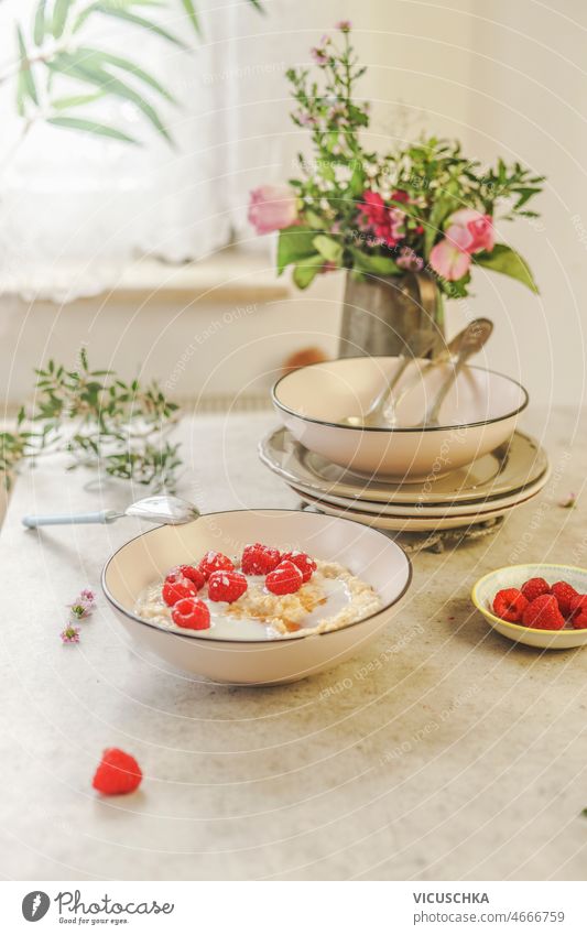 White bowl with vegan porridge with raspberries at white concrete kitchen table crockery flower bunch window background natural light healthy breakfast home