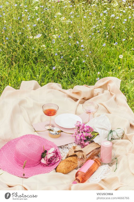 Summer picnic with wine, cheese, baguette, pink sun hat, flowers and candle on beige blanket summer meadow romantic date idea food drink outside front view