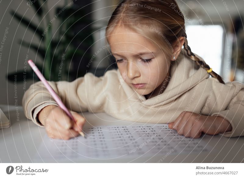 Caucasian kid studying online at home and writing. Pretty serious schoolgirl studying homework math during coronavirus covid-19 quarantine and social distancing
