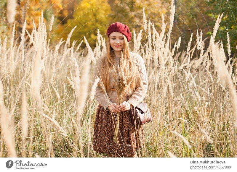 Beautiful woman in autumn park wearing a burgundy beret, beige sweater and brown skirt, standing in dry wheat wild field. Young millennial woman with long hair in stylish fall outfit, smiling and looking to the camera. Autumn female lifestyle, inspiration.