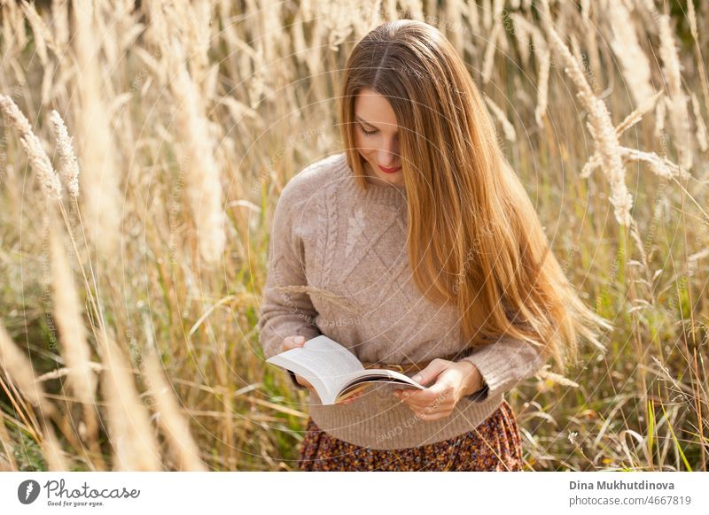 Beautiful woman in reading a book wearing a beige sweater and brown skirt, standing in dry wheat wild field. Young millennial woman with long hair in stylish fall outfit, smiling and looking to the camera. Autumn female lifestyle, inspiration.