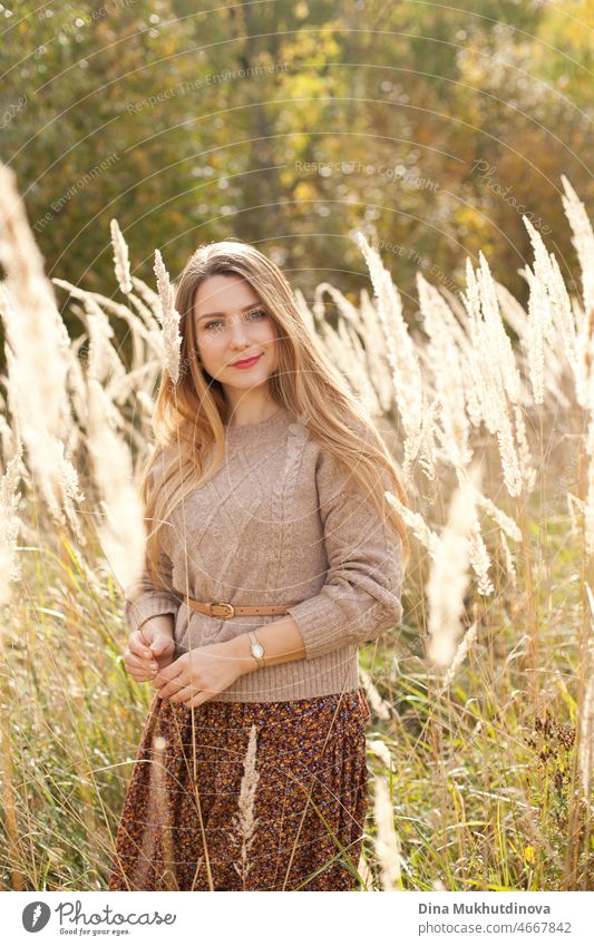 Beautiful woman in autumn park wearing a beige sweater and brown skirt, standing in dry wheat wild field. Young millennial woman with long hair in stylish fall outfit, smiling and looking to the camera. Autumn female lifestyle, inspiration.
