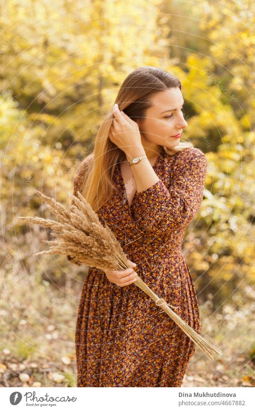Beautiful woman in autumn park wearing a brown dress, holding a dry wheat bouquet. Young millennial woman with long hair in stylish fall outfit, smiling and looking to the side. Autumn lifestyle, inspiration and femininity.