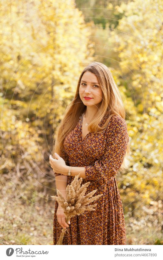 Beautiful woman in autumn park wearing a brown dress, holding a dry wheat bouquet. Young millennial woman with long hair in stylish fall outfit, smiling and looking to the camera.