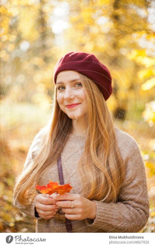 Beautiful woman in autumn park wearing a burgundy beret, holding a red maple leaf. Young woman with long hair in stylish fall outfit, smiling and looking to the camera.