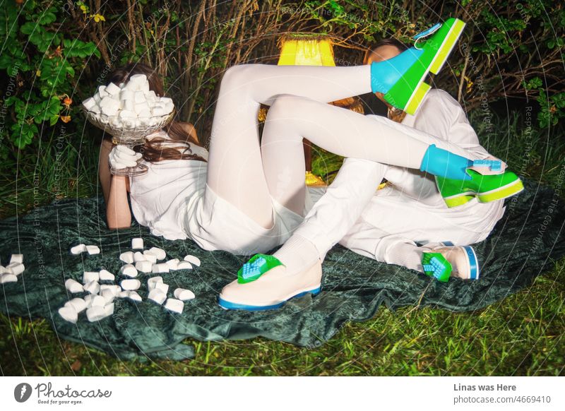 A fashionable couple with some fine white clothes and colorful avant-garde shoes are having delicious marshmallows. Faces aren’t really visible, just the feeling of comfort and flirting in the image. Green bushes are comfy for such a date.