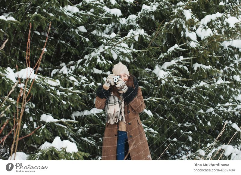 Beautiful woman with long brown hair taking photos on film retro camera walking in winter park with fir trees during snowfall. Winter fashion and stylish outfit. Real people having fun in winter, enjoying fresh air in nature with snowy spruce trees.