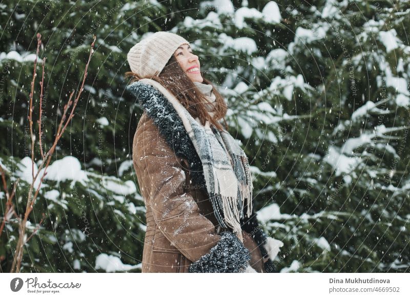 Beautifulwoman with long brown hair walking in winter forest or park with fir trees during snowfall. Winter fashion and stylish outfit. Real people having fun in winter, enjoying fresh air in nature with snowy spruce trees.