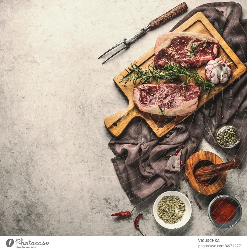 Raw beef steaks preparation with rosemary and garlic at wooden cutting board raw kitchen table dish cloth butcher fork mortar pestle salt pepper chili concrete