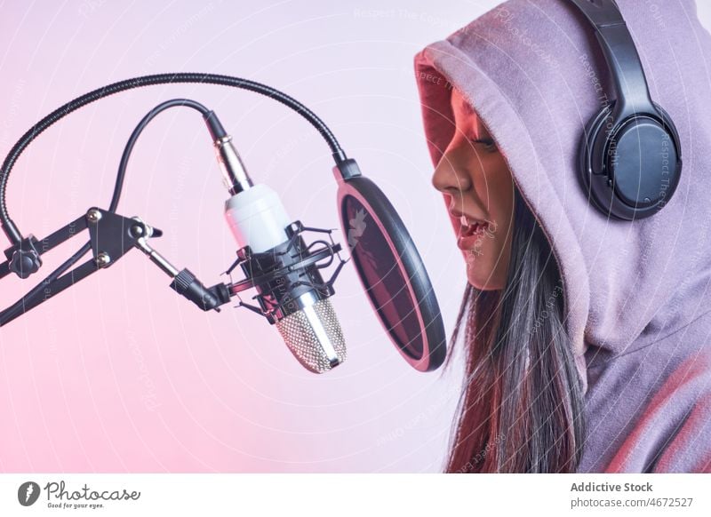 Woman singing into microphone in studio woman singer song vocalist headphones record music voice sound hoodie casual pop filter feminine appearance female young
