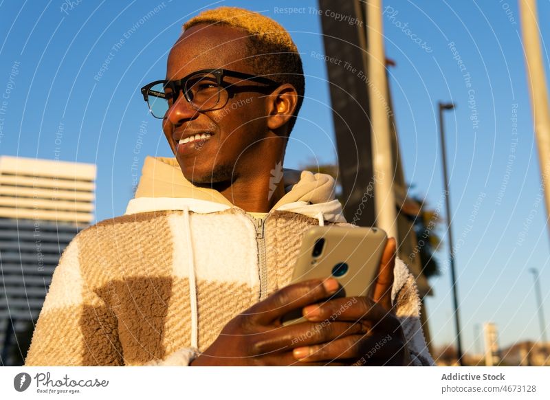 Smiling black man surfing smartphone in city browsing online text message chat digital street building fashion dyed hair modern young short hair male cellphone
