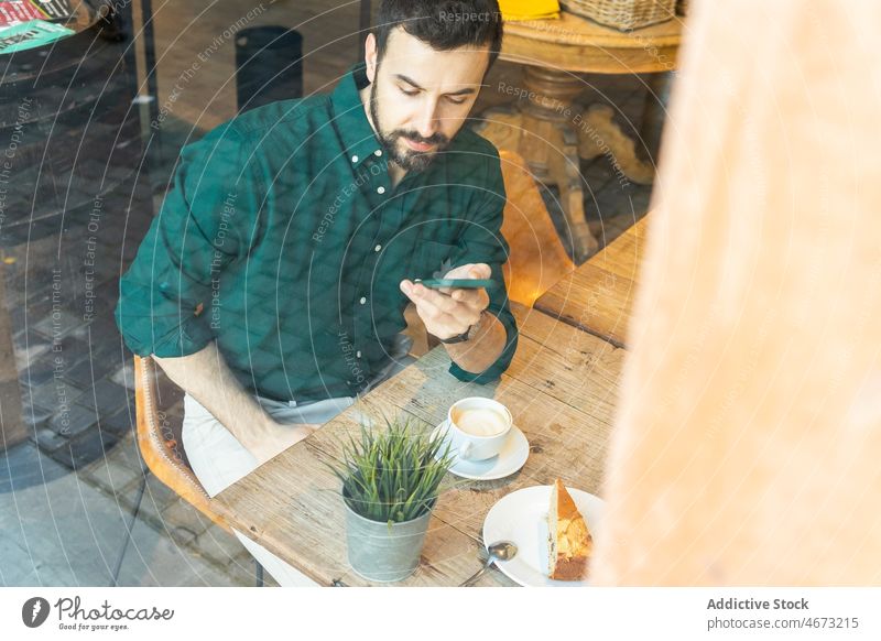 Through glass of man browsing smartphone in cafe online text message coffee entrepreneur break internet hot drink dessert cafeteria table cup delicious beverage