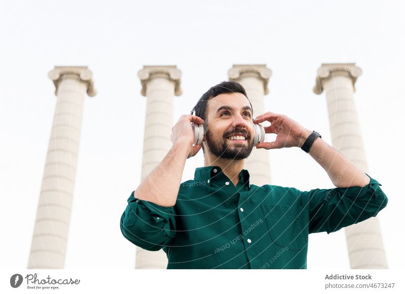 Cheerful man listening to music near columns pillar medieval street ancient headphones song meloman wireless old historic city stone male aged town environment