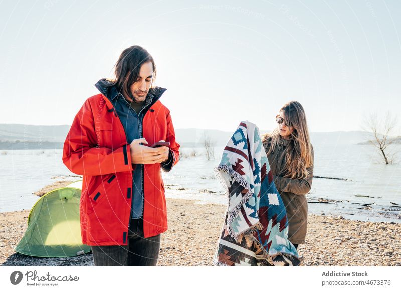 Couple of travelers standing near tent on lake shore couple campsite together autumn mountain pond nature plaid smartphone fall season holiday journey