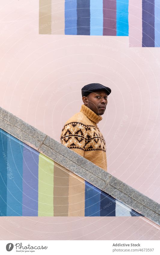Black man in warm sweater walking downstairs warm clothes trendy cap knitted colorful stripe wall casual portrait fashion male outfit style african american
