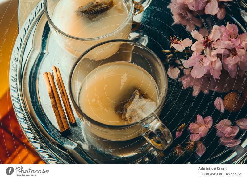 Cups of tea with milk near flowers and cinnamon sticks hot drink beverage branch serve teatime floral plant cup glass tray twig blossom tasty natural table room