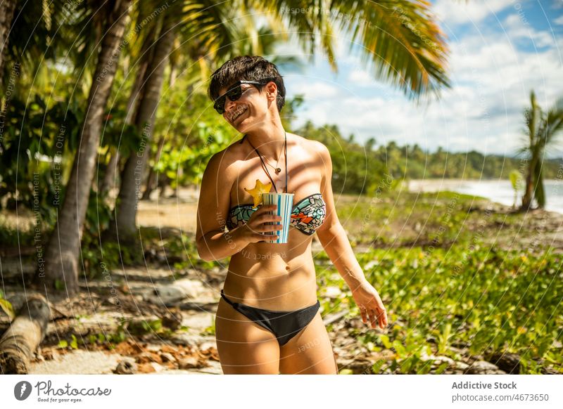 Positive woman in bikini with beverage on coast swimwear refreshment shore recreation nature sea tropical tree rest tourist pastime trip water female summer