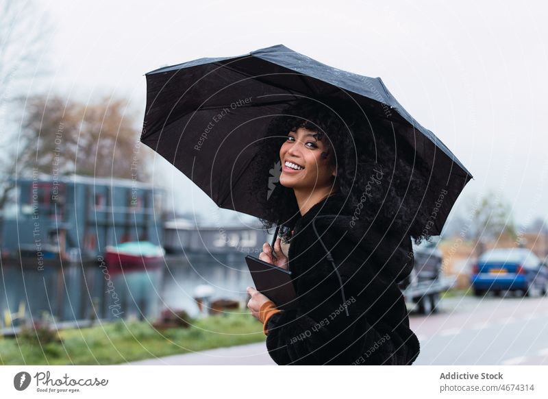 Smiling black woman with umbrella on street rain city roadside urban appearance tablet female portrait african american woman casual young happy positive smile