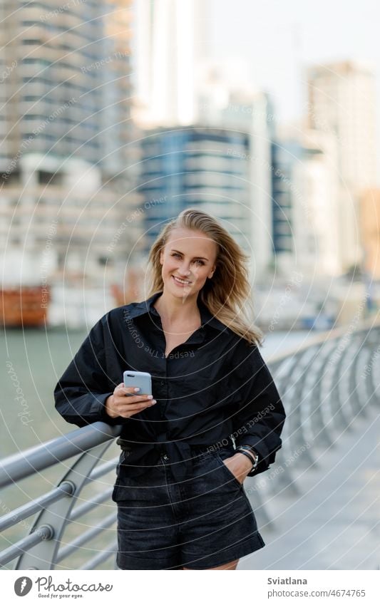 An attractive woman stands on the embankment and uses her phone while walking around the city blonde street stylish beautiful vacation smiling using traveller