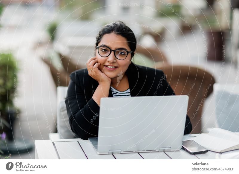 A young and confident Indian Asian woman smiles while sitting and working on her laptop in a stylish cafe on the street on a summer day. girl indian asian