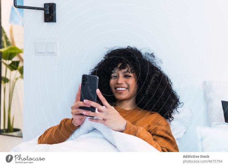 Happy black woman using smartphone in bed awake morning weekend at home relax rest female african american mobile browsing internet smile happy young curly hair
