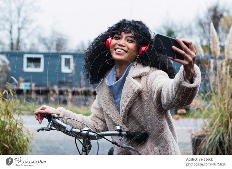 Delighted black woman with smartphone on bicycle headphones using music taking selfie listen ride bike chill female african american woman relax city portrait