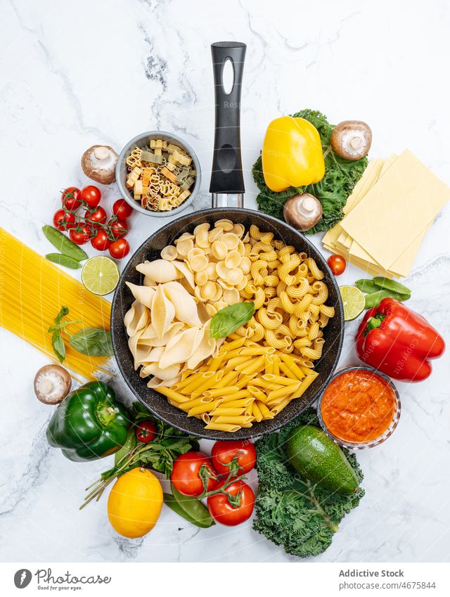 Frying pan with assorted raw pasta amidst vegetables various frying pan uncooked kitchen food recipe culinary cuisine ingredient product prepare bell pepper