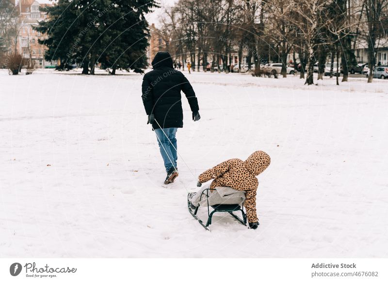 Anonymous father pulling cheerful kid on sled fatherhood childhood winter snow amusement having fun play street season frost outerwear cold wintertime man dad