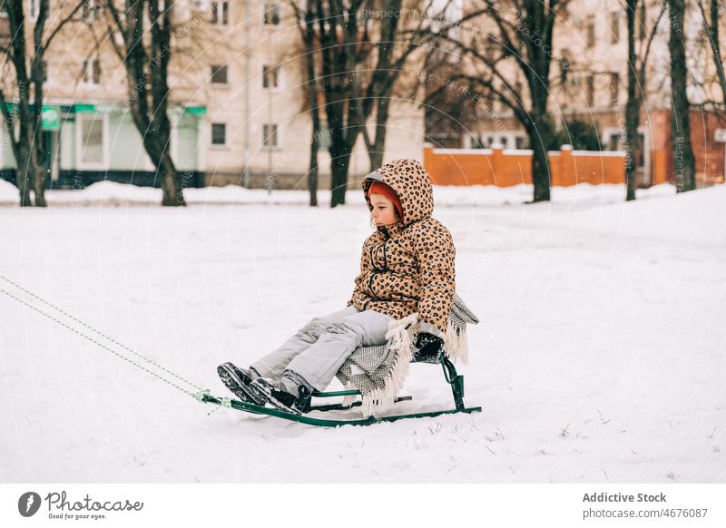 Kid sitting on sled in winter day kid childhood snow street city season frost outerwear cold weather warm clothes temperature frozen activity active equipment