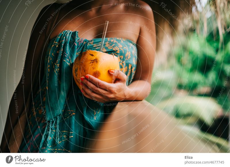 Woman wrapped in colorful bath towel holds a King coconut with straw in hand. Vacation in the tropics. Coconut Drinking coconut vacation Summer Tropical