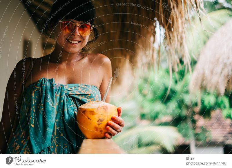 Smiling, colorful bath towel wrapped, woman with sunglasses holds a King coconut with straw in her hand. Vacation in the tropics. Coconut Drinking coconut Woman