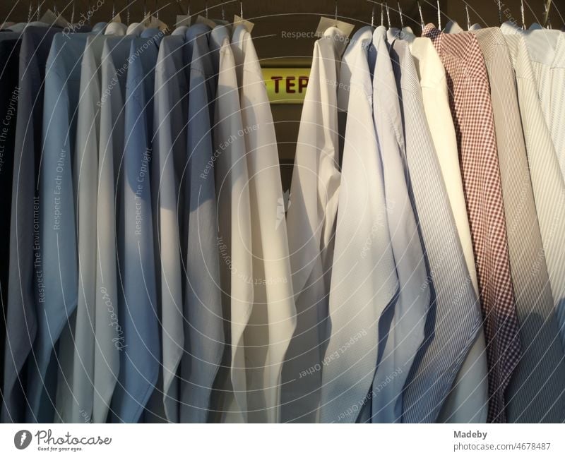 Freshly washed and ironed monochrome men's shirts in natural colors and pastel shades behind the shop window in the sunshine in the district of Sachsenhausen in Frankfurt am Main in the German state of Hesse