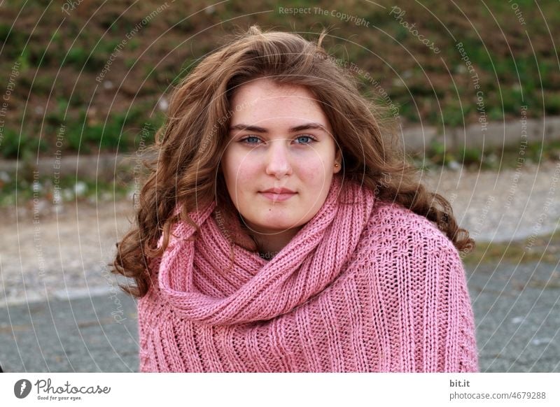 Pretty young curly haired woman wooly dressed looking at camera. Teenager, teenager in nature smiles contentedly in autumn. Beauty photography in pink poncho, coat, jacket made of wool. Beautiful lady with eye contact in front of meadow, lawn, grass, road, asphalt.