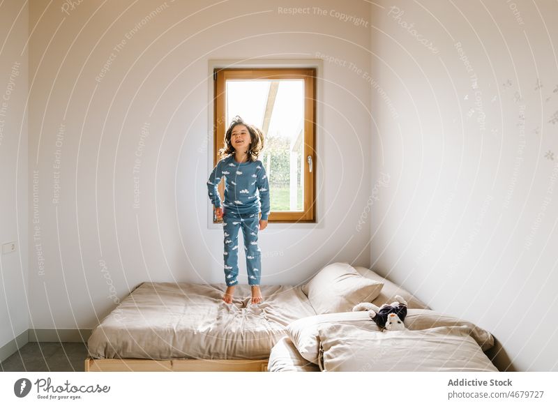 Cheerful girl in pajama jumping on bed kid window bedroom childhood morning having fun activity active apartment playful domestic adorable flat cute home