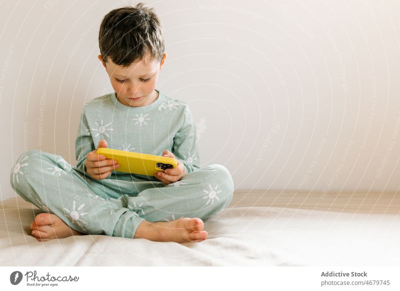 Boy in pajama playing on smartphone boy kid childhood bed bedroom videogame pastime morning domestic apartment adorable barefoot flat cute home residential