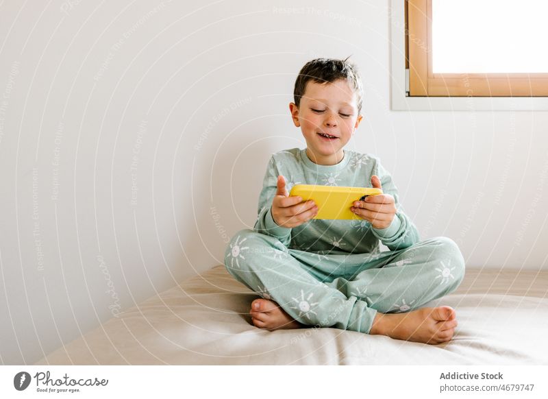 Cheerful boy in pajama playing on smartphone kid childhood bed bedroom videogame pastime morning domestic apartment adorable barefoot flat cute home residential
