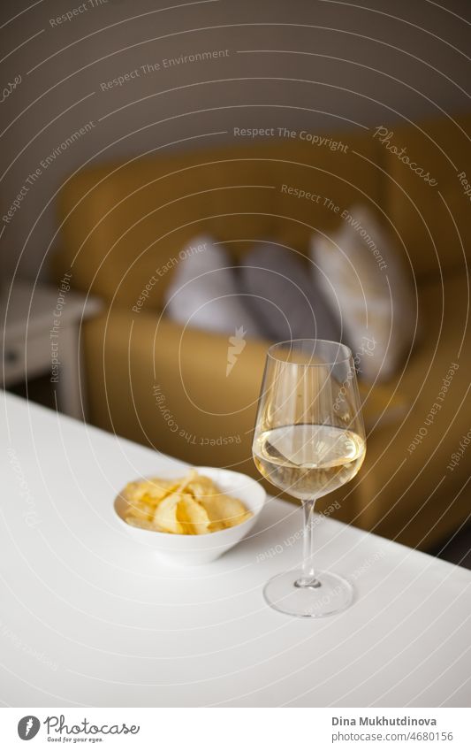 Single white wine glass and potato chips on white table at home. Single apartment living. Home interior with yellow couch. Relaxing evening alone at home. One-person households are becoming increasingly common across the world. Millennial lifestyle.
