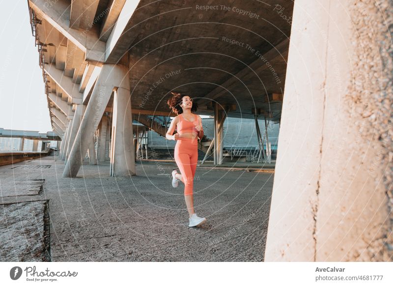 Young multi cultural woman smiling to camera while jogging on urban city to lose weight before summer to get a defined body. Running and workout outdoors. Sunset scene with young athletes