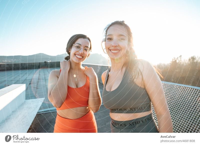 Two young multi cultural woman smiling to camera before starting to train to lose weight before summer to get a defined body. Running and workout outdoors. Sunset scene with young athletes