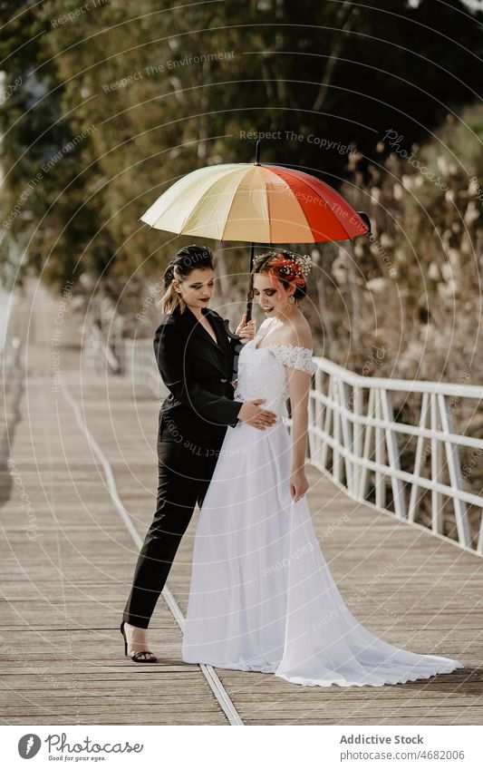 Lesbian couple hugging on bridge during wedding women bride park love umbrella lesbian female together newlywed elegant suit white dress embrace weather