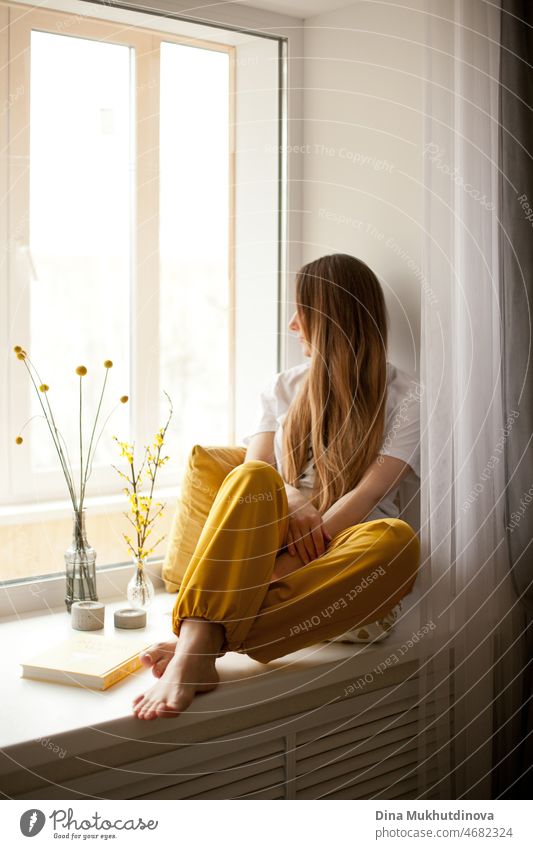 Young beautiful woman wearing white t-shirt and yellow sweatpants reading a book at home, sitting on windowsill, smiling. Cozy relaxed casual lifestyle full-body portrait. Literature and fiction books.  Cozy home room.