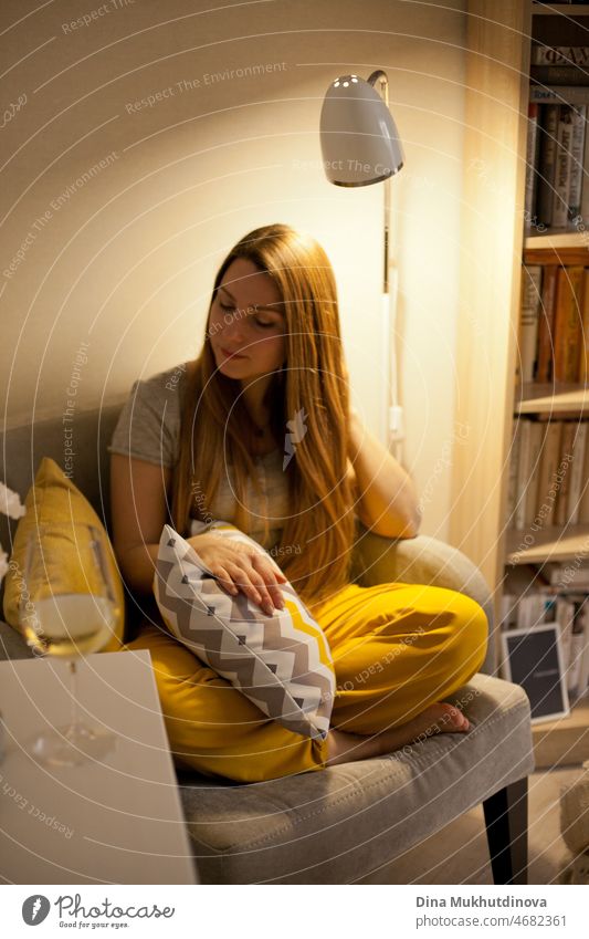 Young beautiful woman wearing gray and yellow sweatpants, sitting in cozy chair, holding a pillow. Literature and fiction books.  Cozy home room. people reading