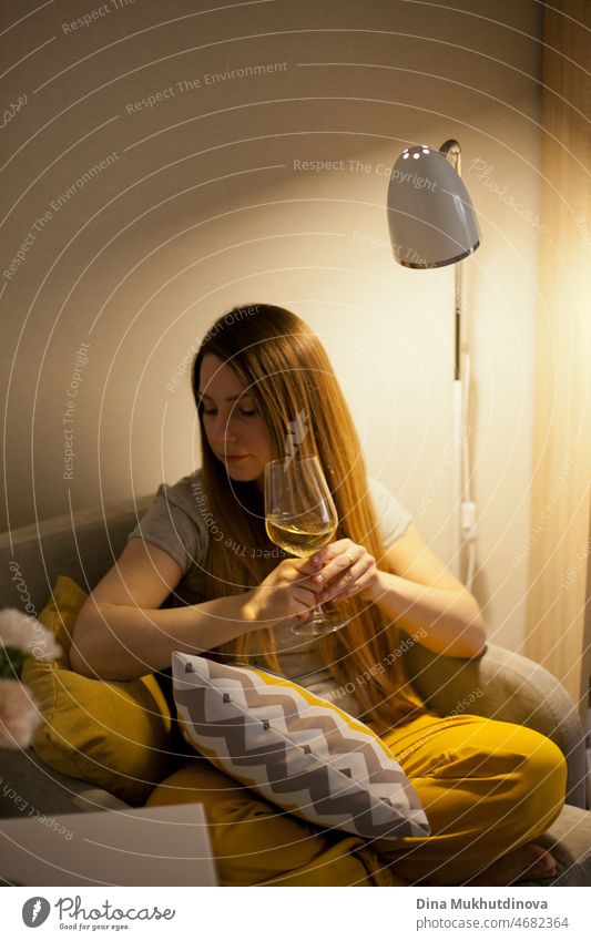 Young attractive woman sitting at home in coxy chair drinking white wine. Treat yourself after hard day at work. Tasting wine. Yellow color in home interior. Casual everyday millennial lifestyle.