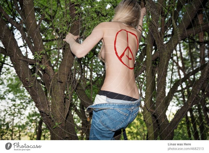 Such a lustful view in this deep forest. A topless blonde girl climbing up in a tree with a devil’s sign on her sexy back. Blue jeans and black lingerie is all that she is wearing.